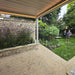 Outdoor patio with stone wall, camel coloured pergola, garden, and people in the background