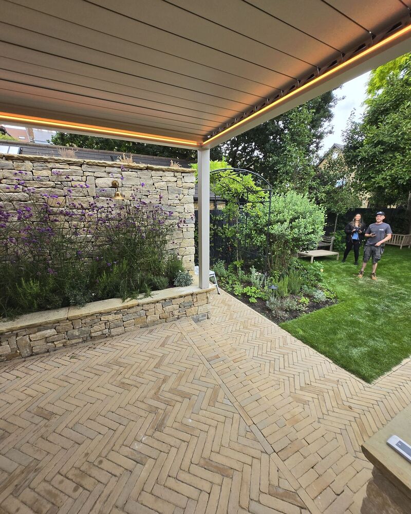 Outdoor patio with stone wall, camel coloured pergola, garden, and people in the background