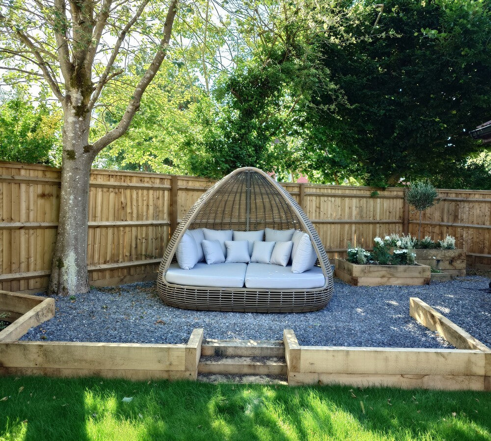 Garden setting with a circular wicker seat and cushions on a gravel area, surrounded by trees and plants.
