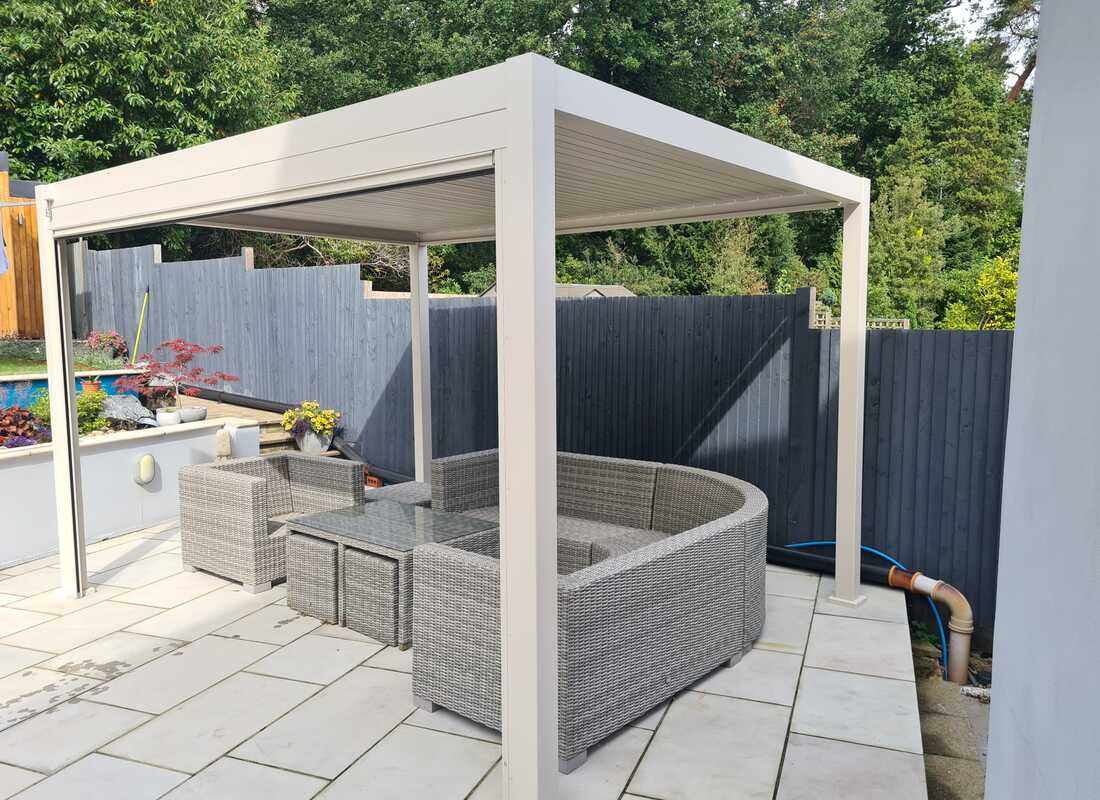 Patio with outdoor furniture under a white pergola, surrounded by greenery.